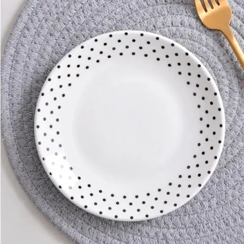 Tallerken fra middagsservice, hvit med svarte prikker langs kanten, sett ovenfra på grått underlag med gylden gaffel.