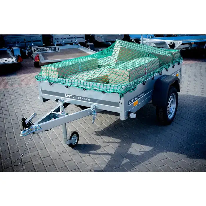 Sikkerhetsnett for tilhenger, grønt nett spent over esker i grå tilhenger på brostein med støttehjul.