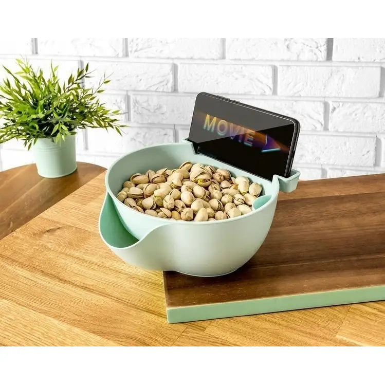 Smart skål med dryppebrett i lys grønn plast, fylt med pistasjnøtter, sett skrått med synlig mobilholder.