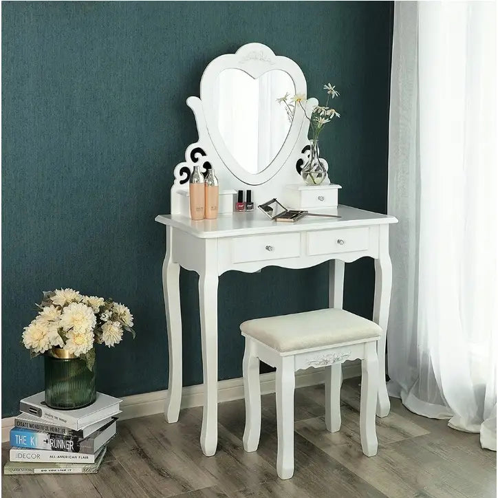 Sminkebord med hjerteformet speil, hvit krakk, buede ben, to skuffer og sminkeprodukter på bordet.