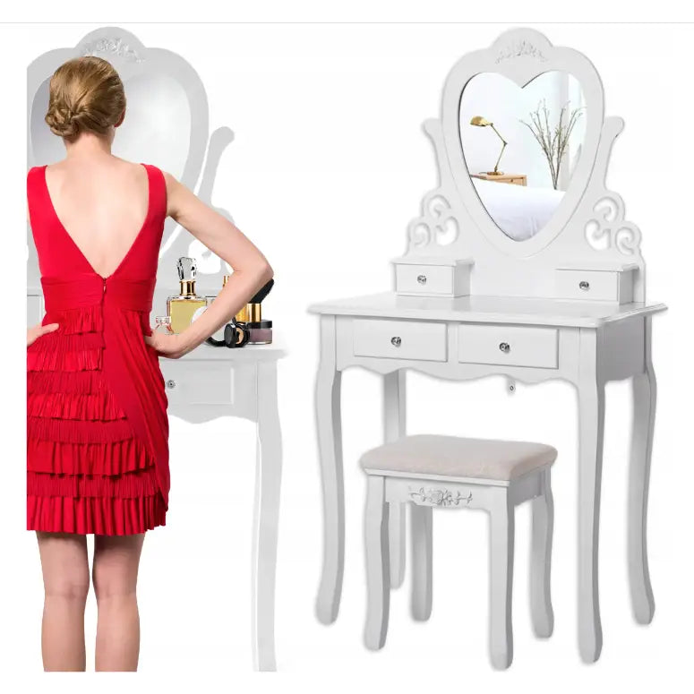 Sminkebord med hjerteformet speil, hvit krakk og kvinne i rød kjole som sminker seg ved bordet.