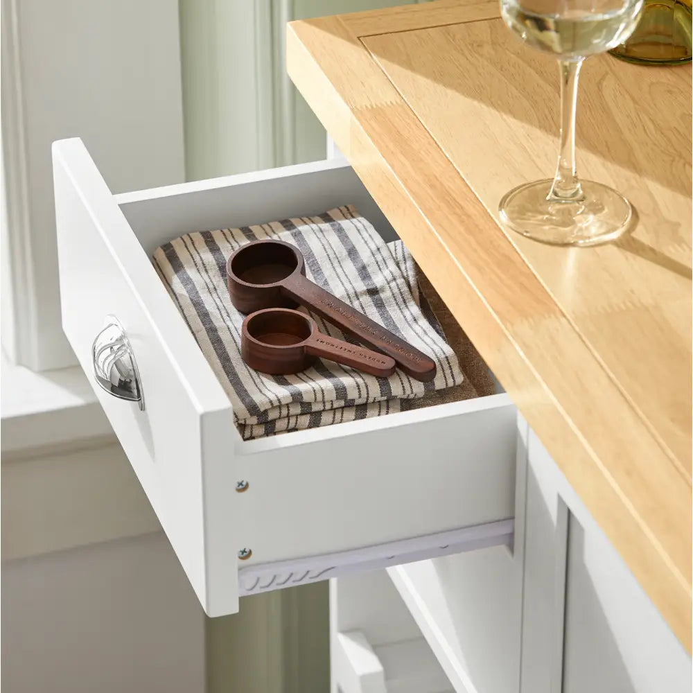 Sobuy kjøkkenøy, hvit skuff trukket ut med stripete håndkle og tre måleskjeer, lys treplate og vinglass.