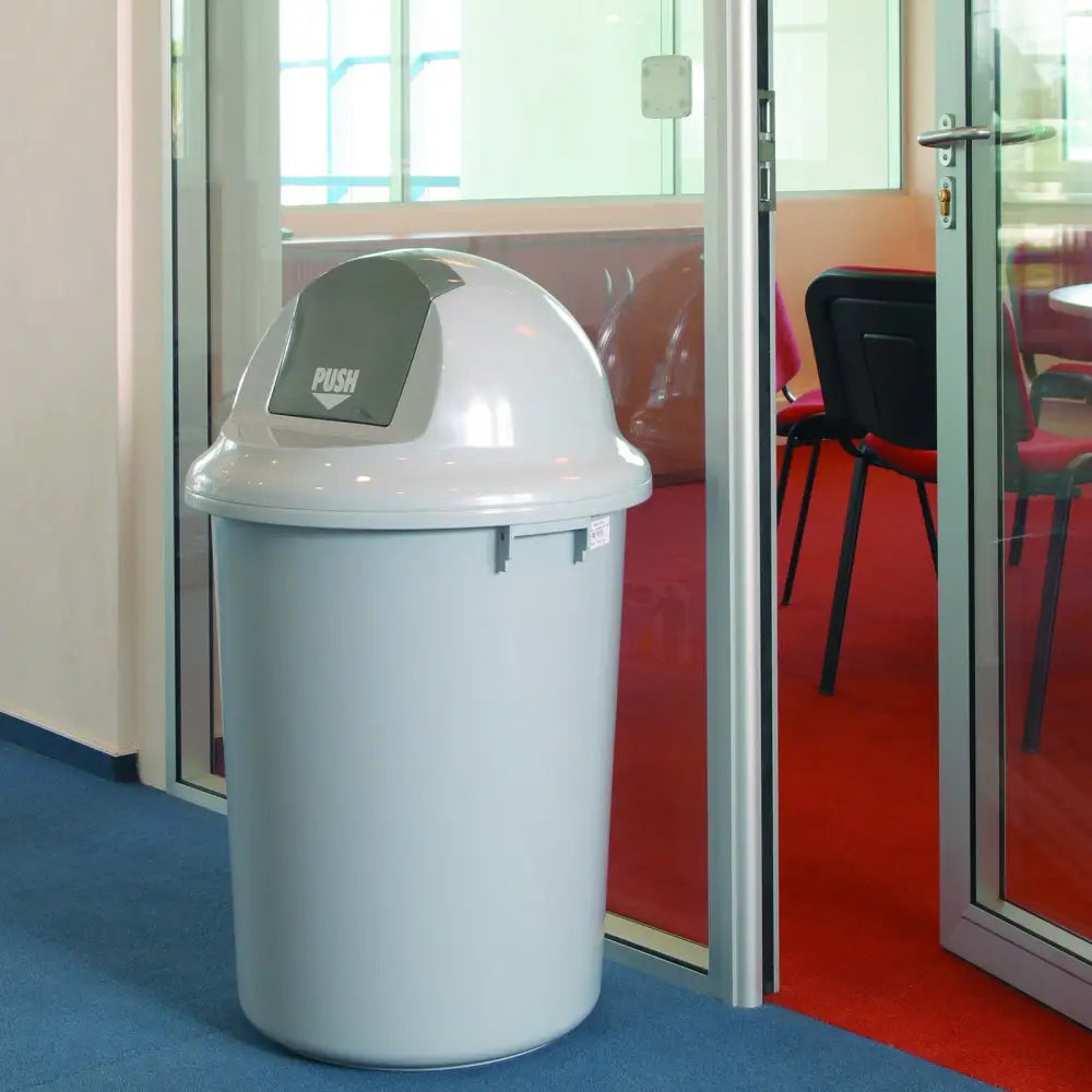 Søppelbøtte med skrå klaff PUSH i grå plast, vist ved glassdør i moderne kontormiljø.