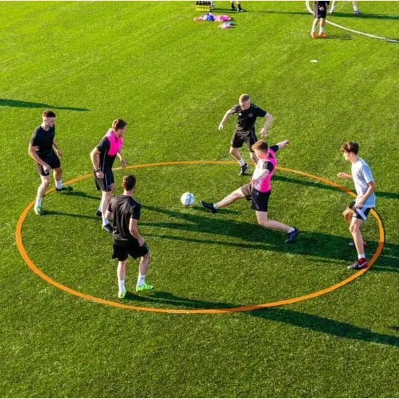 Spillet hjul Tiki Taka, seks personer spiller fotball i oransje sirkel på grønn kunstgressbane, sett ovenfra.