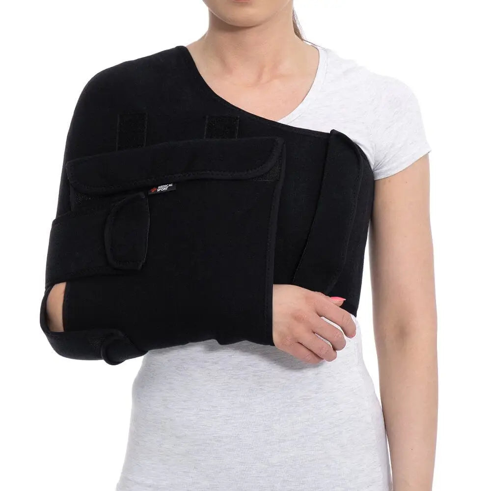 Stabilisator skulder- og armortose i svart stoff, sett forfra på person med hvit T-skjorte og justerbare stropper.