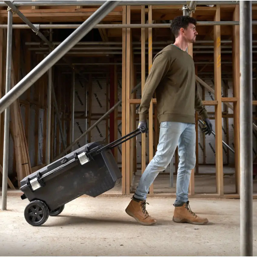 Stanley verktøykasse på hjul, trilles av mann med hansker på byggeplass med trekonstruksjoner.
