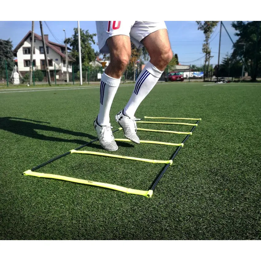 Stige Stige agilitystige, sett i lav vinkel mens en utøver trener på kunstgress med hvite fotballsko.