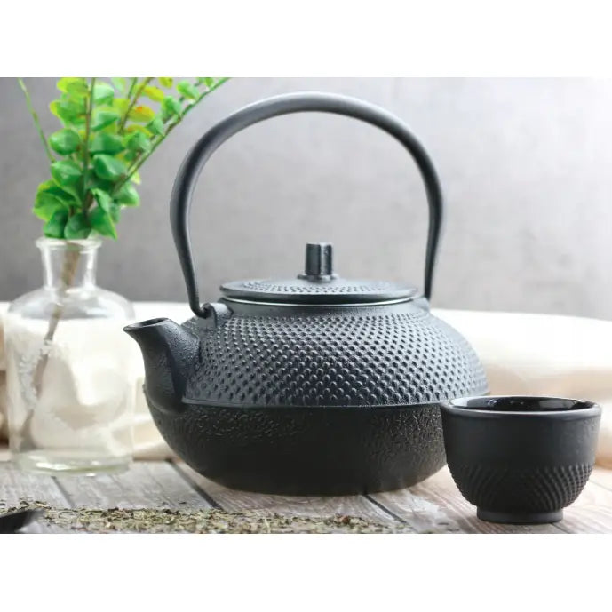 Støpejern tekanne med infuser, teksturert sort overflate, plassert ved siden av kopp på trebord.