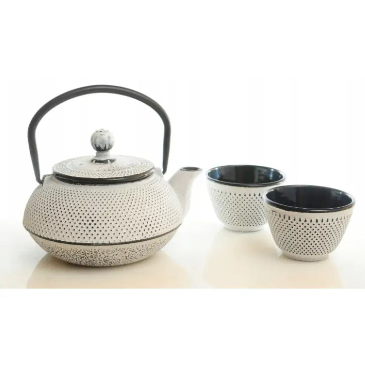 Støpejernskanne med prikkete hvit overflate, sort håndtak og to kopper med sort innside, sett fra siden.