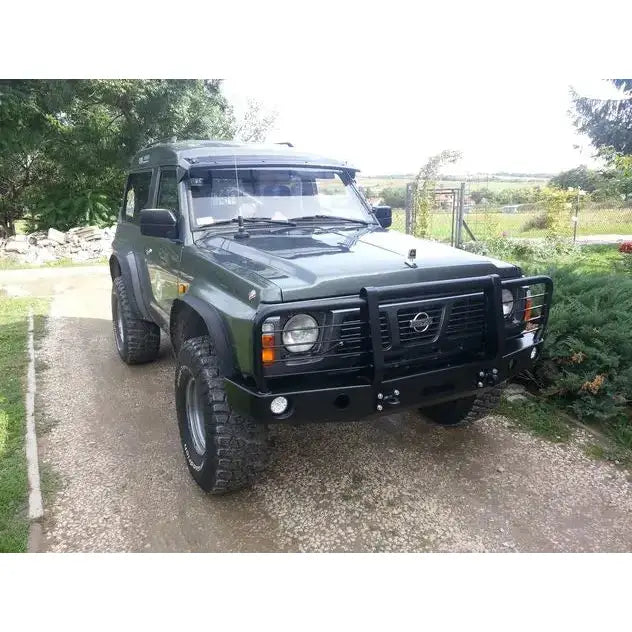 Front bumper på Nissan Patrol Y60 med sort bullbar, ekstra lys og grove terrengdekk på grusvei.