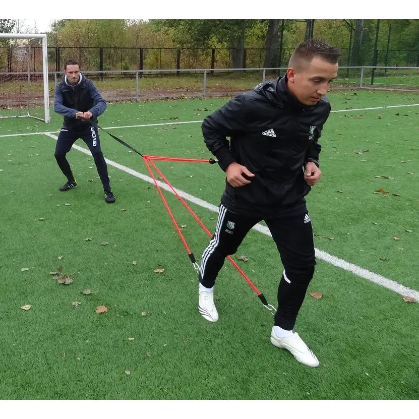 Styrketreningsgummi Edderkopp i bruk, to personer koblet sammen med røde strikker under løpstrening på fotballbane.