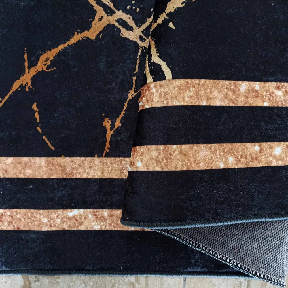 Svart og gullfarget teppe med marmormønster, brede gullstriper og brettet hjørne som viser undersiden.