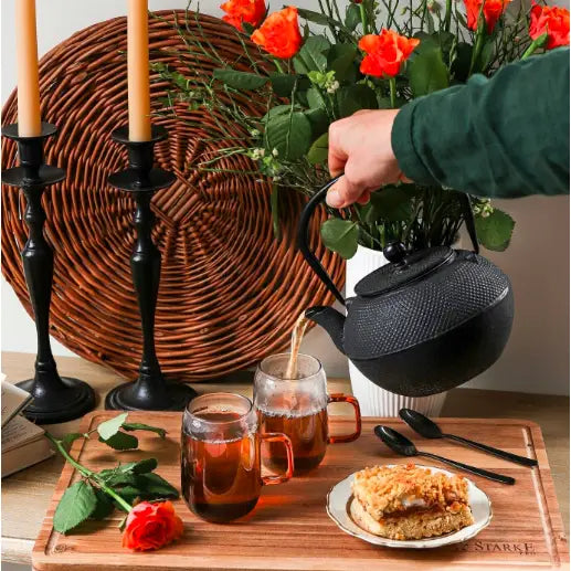 Svart tekanne i støpejern med tefilter, holdt over glass te på brett med rose, bestikk og kake.