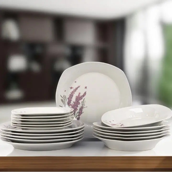 Porselenssett med hvite tallerkener, lilla blomsterdekor, stablet i hauger på hvit overflate i lyst rom.