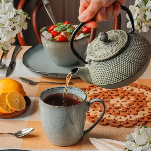 Tekanne Konighoffer 0,8L i grønt støpejern, hellende te i grå kopp, dekorert med blomster og sitroner.