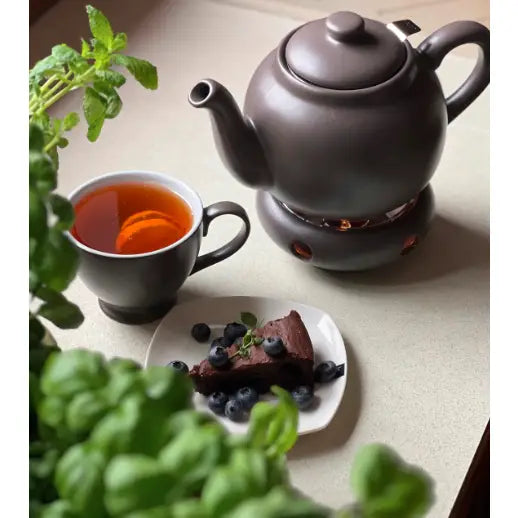 Tekanne med infuser i grått steintøy, sett skrått ovenfra med kopp te, sjokoladekake og friske urter.