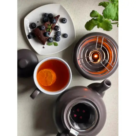 Tekanne med infuser i mørk grått steintøy, sett ovenfra med kopp, appelsinskive i teen og blåbærkake ved siden av.
