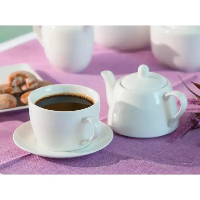 Tekanne med kopp i hvitt porselen, sett skrått på lilla duk med mørk drikk og uskarpe kjeks i bakgrunnen.