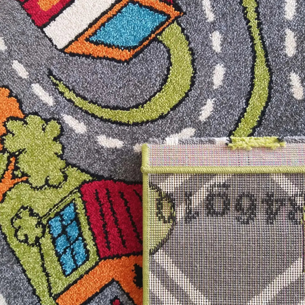 Tepe Bella grå barneteppe med veibanemønster, hus og trær, detalj av brettet hjørne og synlig kanting.