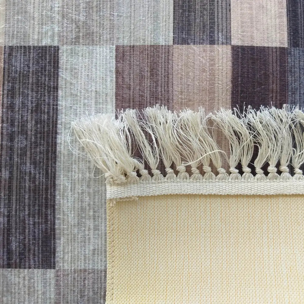 Tepe Blanka brun teppe, detalj av hjørne med beige frynser og synlig bakside mot rutete bakgrunn.