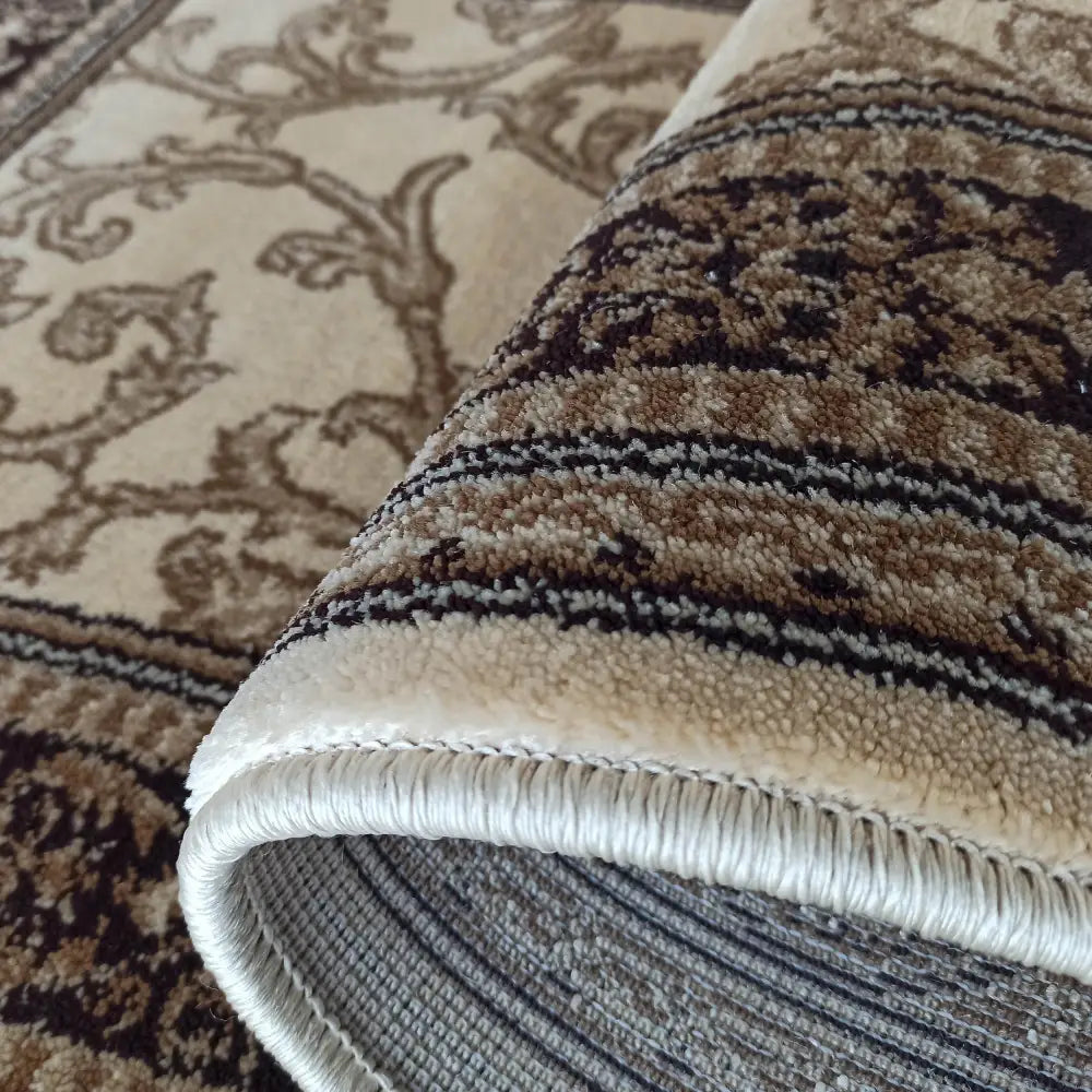 Tepe Eksklusiv teppe i krem, nærbilde av tykk kanting og klassiske beige og brune mønstre, delvis rullet opp.