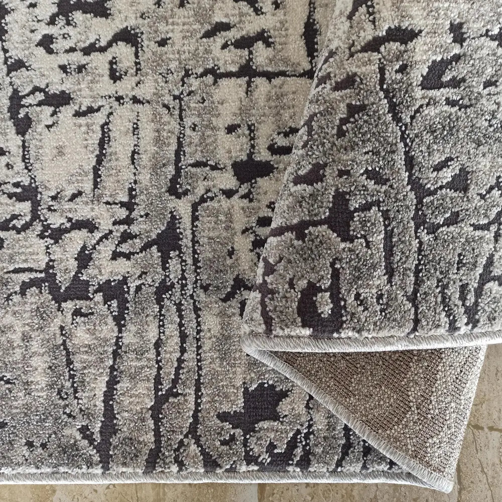 Tepe Enigma teppe, nærbilde av grått og sort abstrakt mønster, oppbrettet hjørne viser kant og underside.