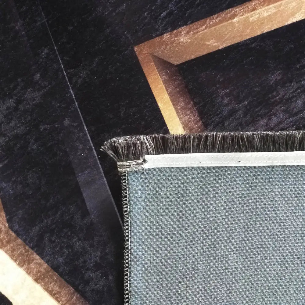 Tepe i svart og gull, nærbilde av hjørne med grå bakside og dekorativ kant mot et mørkt mønstret teppe.