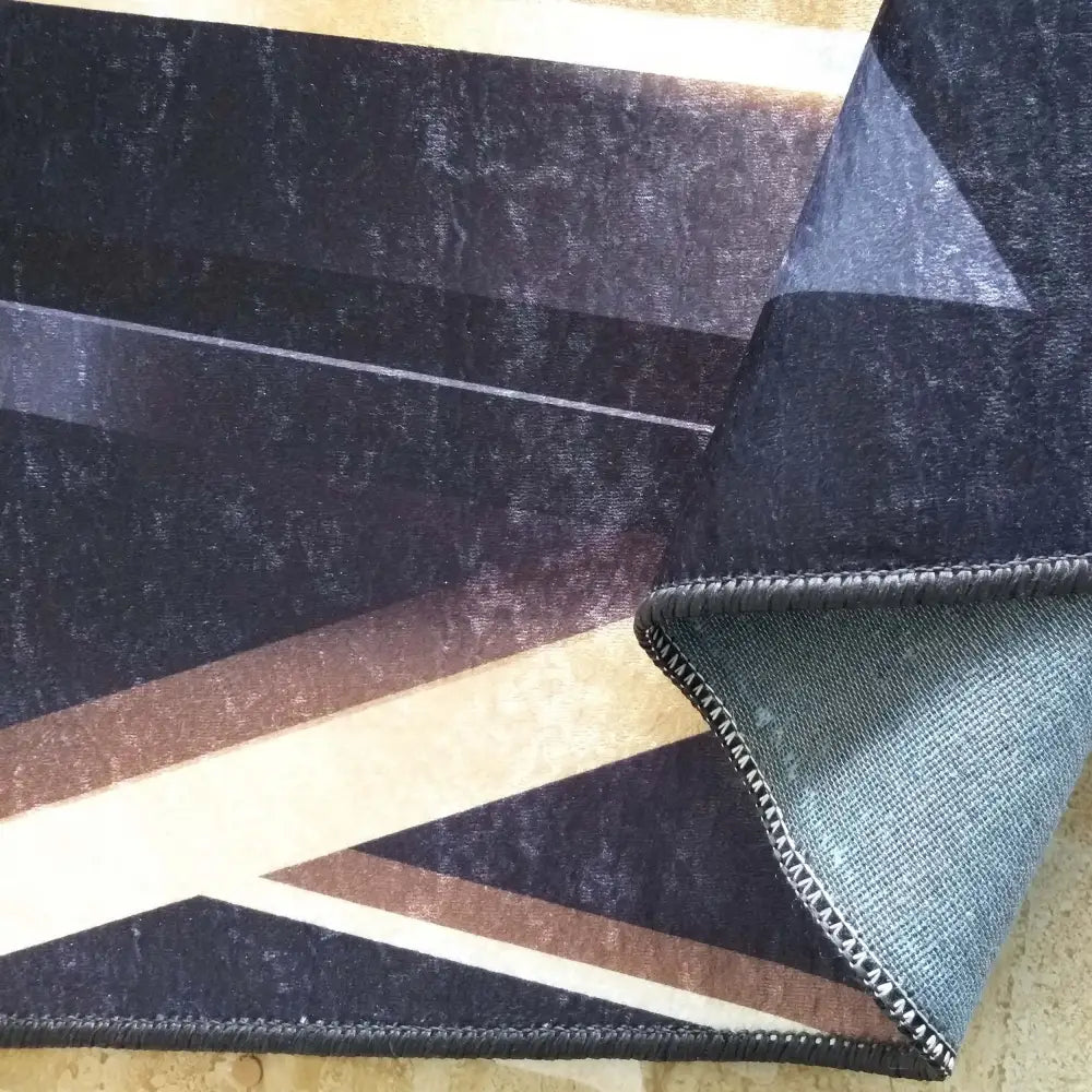 Tepe i svart og gull, nærbilde av geometrisk mønster med oppbrettet hjørne som viser baksiden og sømkantene.