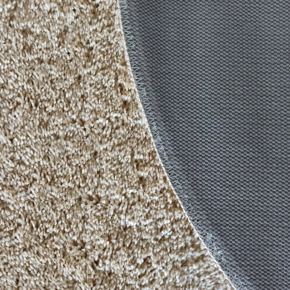 Tepe Kamel Beige rundt teppe, detalj av kanten med myk beige luv og grått antiskli-materiale under.