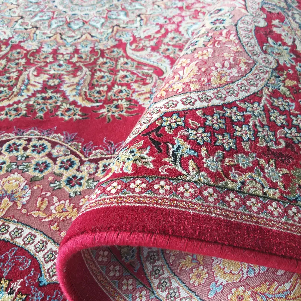 Tepe luksuriøst teppe i røde og rosa nyanser, detaljert hjørne viser tykkelse og blomstermønster.