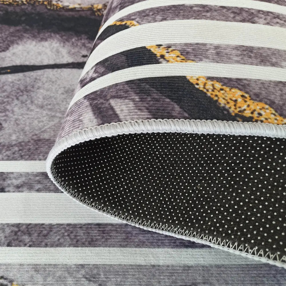 Tepe mørk grå teppe, nærbilde av stripet overflate med gull, hvitt, antiskli-prikker og overlock-søm.