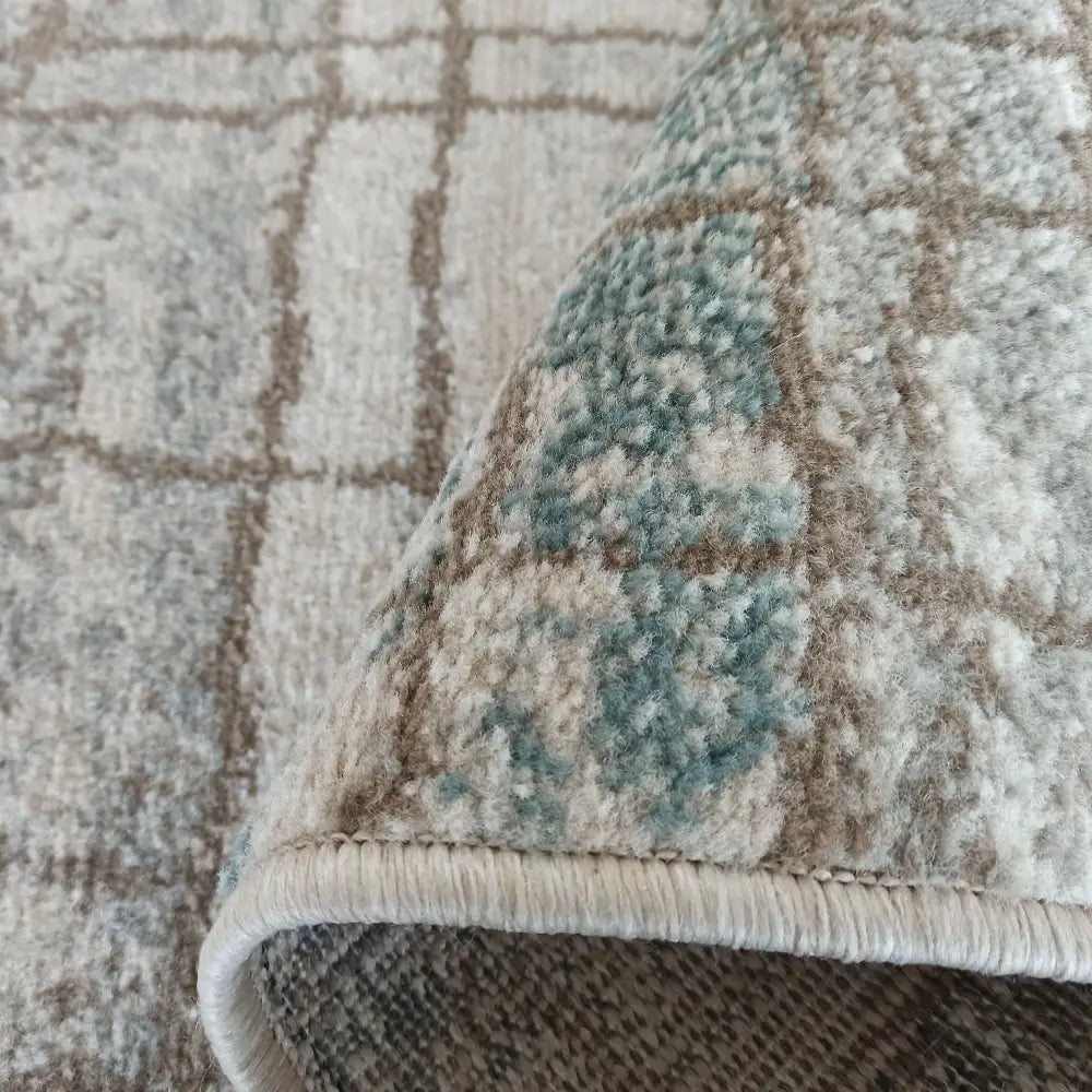 Tepe Roxanne teppe, nærbilde av oppbrettet hjørne med blå, beige og brune ruter og synlig tekstur.