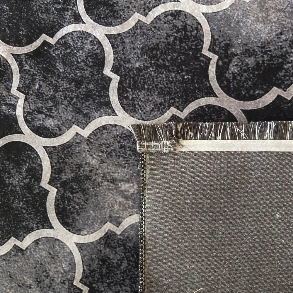 Tepe svart teppe 160 x 220 cm, nærbilde av grå bakside, kant med frynser og sort-hvitt geometrisk mønster.