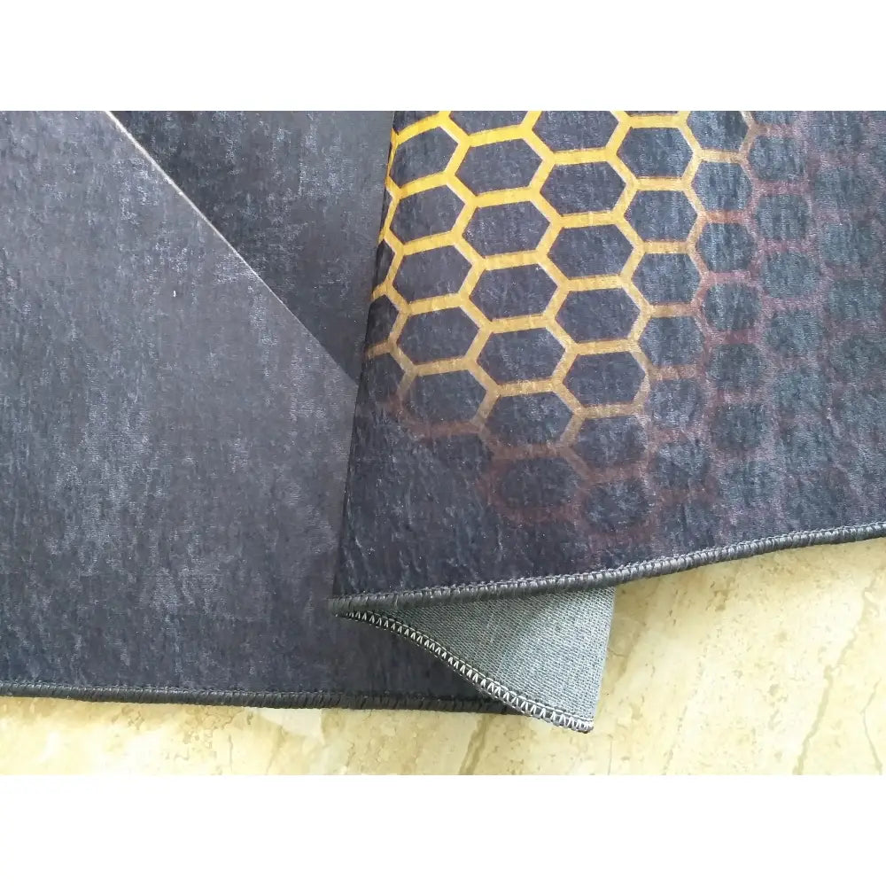 Tepe svart og gull håndkle, nærbilde av sekskantmønster, synlig søm og brettet hjørne som viser tekstur.