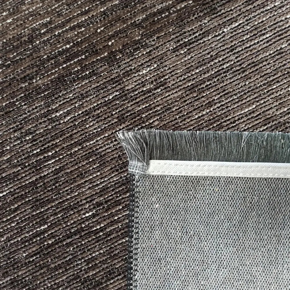 Tepedrøm Diamond 02 brun, nærbilde av hjørne med grå frynser og synlig tekstur mot mørkt mønstret teppe.