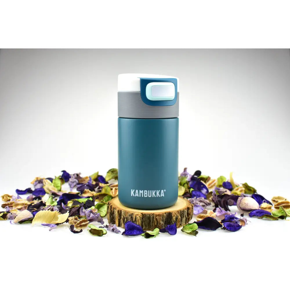 Termokopp Kambukka Etna 300ml i dyp turkis med grått og hvitt lokk, plassert på treflis med tørkede blomsterblader.