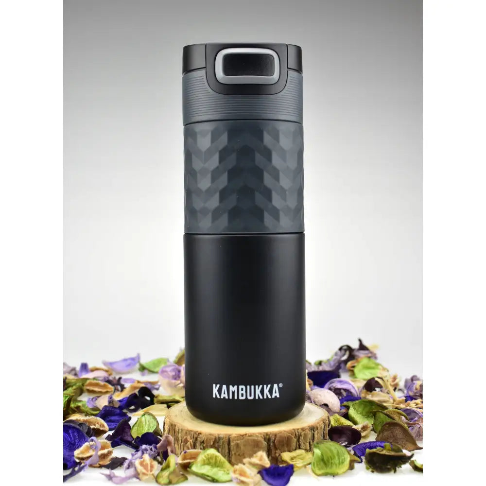 Termokopp Kambukka Etna Grip 500ml i svart stål med grått lokk, plassert på trestamme omringet av tørkede blomster.