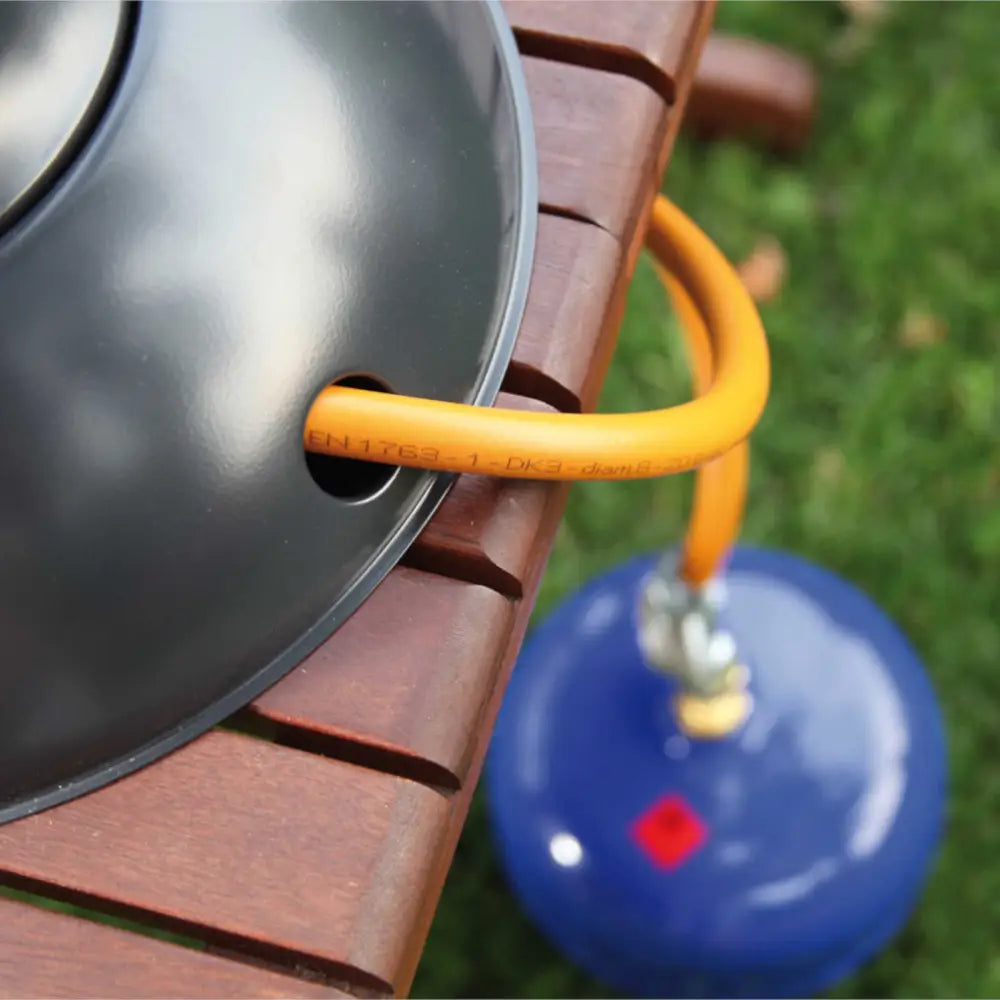 Terrassevarmer ETNA, nærbilde av svart metallskive og oransje gasslange koblet til blå beholder på trebenk.