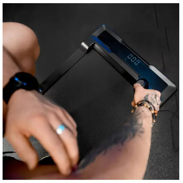 Sort digital vekt med LED-display holdt i hånden av person med tatovert arm, sett ovenfra mot mørk bakgrunn.