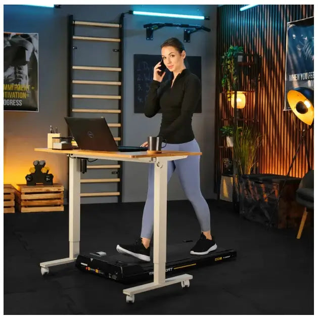 Tredemølle for hjemmetrening AERO Trex Sport under skrivebord, kvinne i gangtempo i moderne treningsrom.