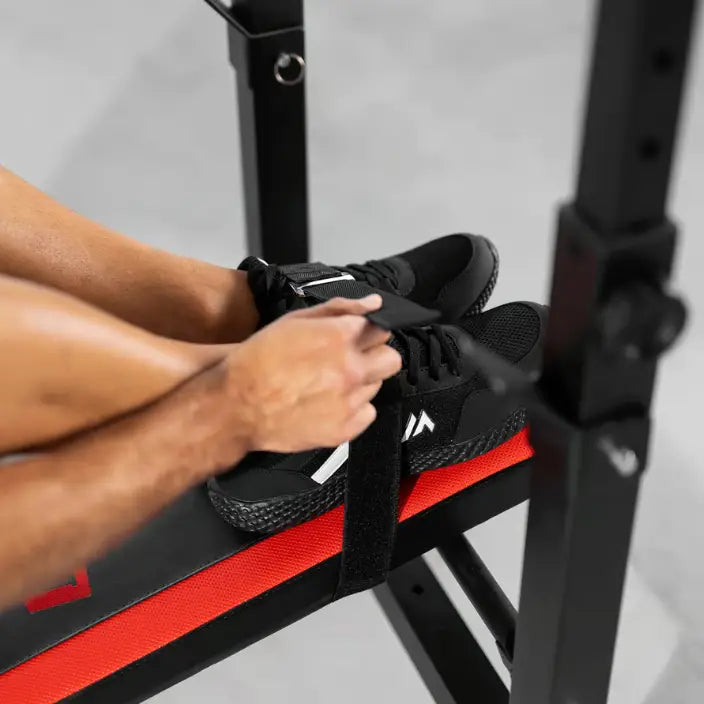 Treningsbenk med vektstangsett, nærbilde av føtter med borrelåsfeste og hender som justerer reim på rødt trekk.