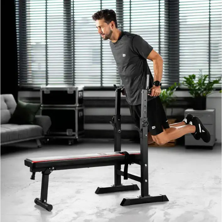 Treningsbenk med vektstangsett, sort med røde detaljer, mann viser dips i hjemmegym.