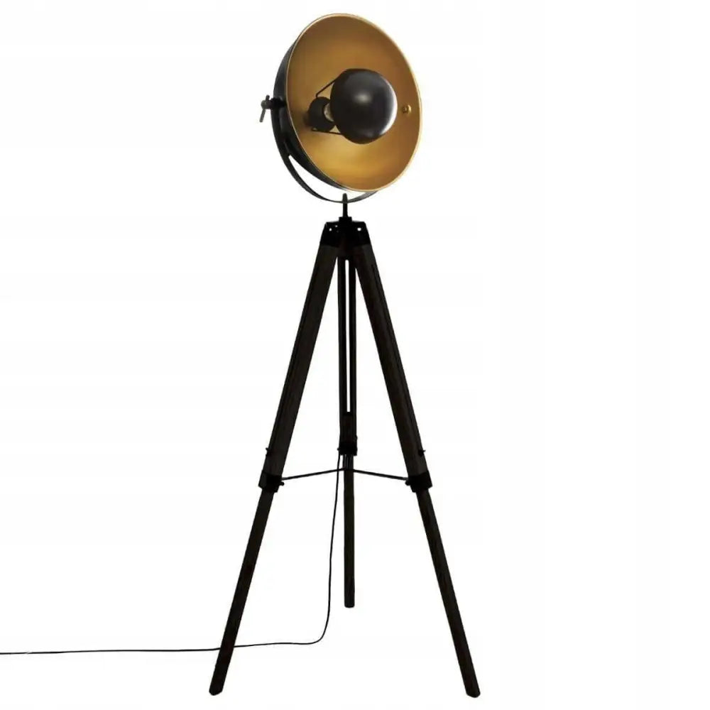 Tripod gulvlampe med svart trefot, stor gyllen skjerm og synlig ledning, sett fra siden mot hvit bakgrunn.