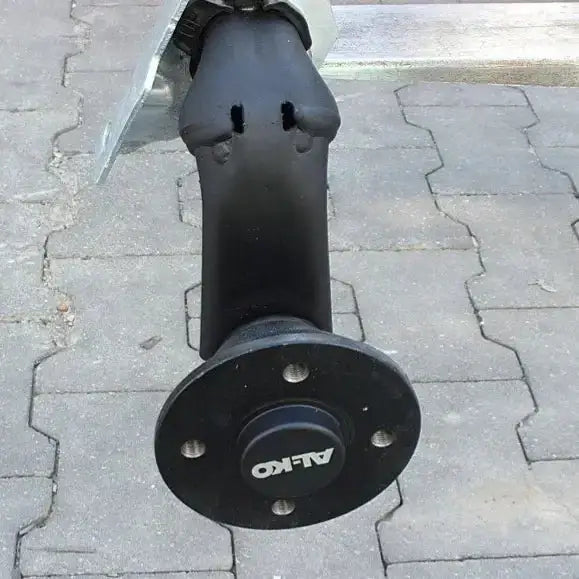 Aksling tilhenger AL-KO, frontvisning av sort metallaksel med hjulnav og tydelig AL-KO-logo på belegningsstein.