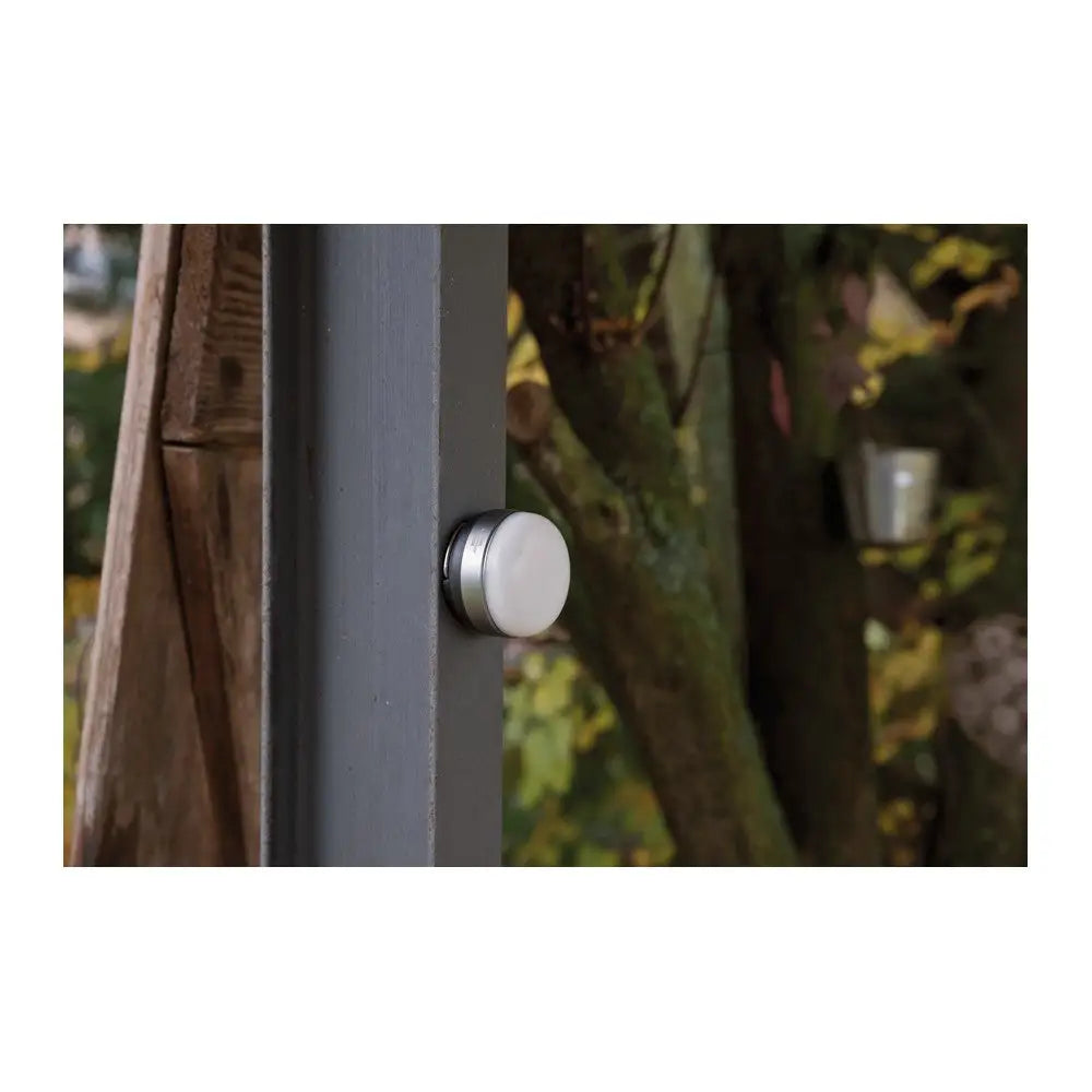 Utendørslampe LED OLI, rund hvit sensor med sølvkant montert på mørk stolpe ute blant trær.