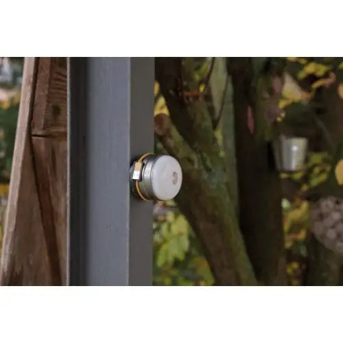 Utendørslampe LED med rund hvit sensor, svarte og gull detaljer, montert på grå stolpe blant trær.