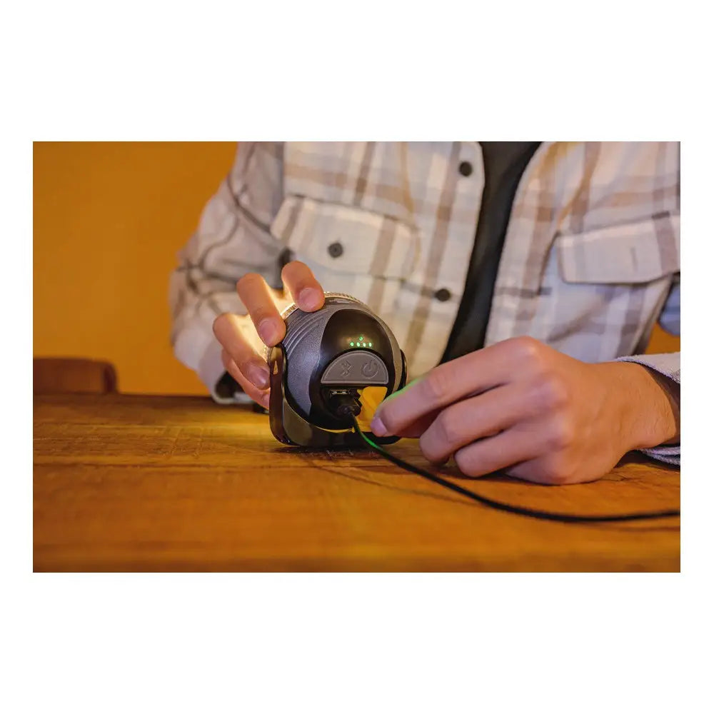 Utendørslampe LED OLI 310 AB med høyttaler, bakside med ladeport og knapper, holdt i hånden på trebord.