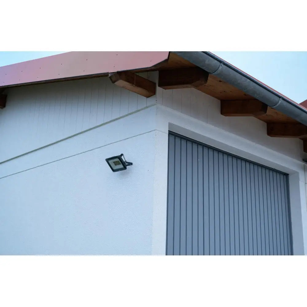 LED vegglampe, svart rektangulær modell montert på lys murvegg ved hjørnet med garasjeport og tretakbjelker.