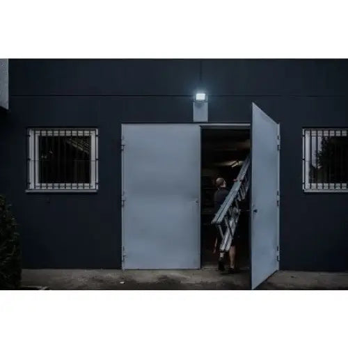 Vegglampe LED JARO 7060 P montert over grå metalldør, lyser opp inngang med person og stige.