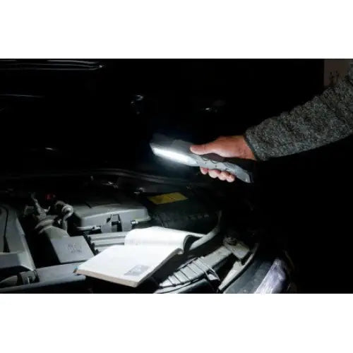 Verkstedslampe LED, holdt over bilmotor i mørket, lyser opp motordeler og åpen instruksjonsbok.